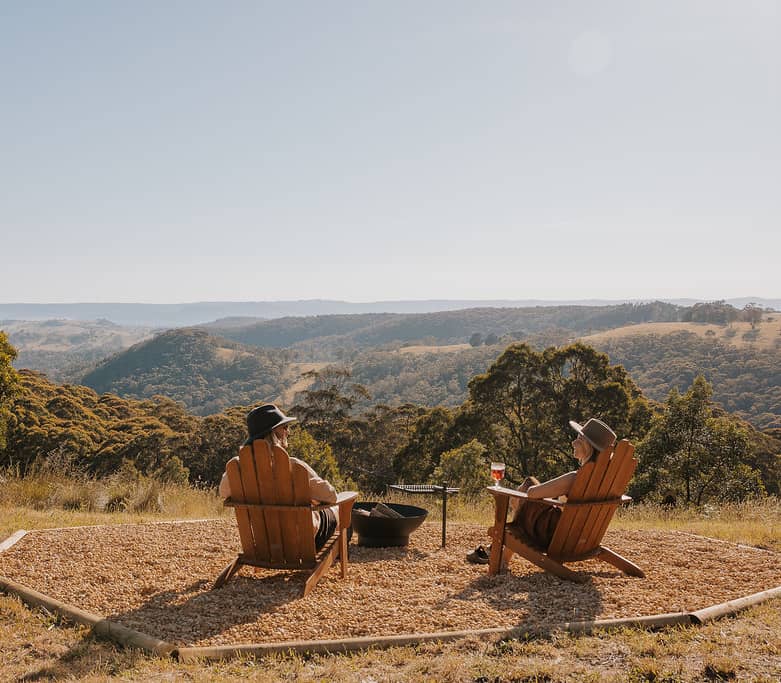 A couple sits in wooden chairs by a fire pit overlooking a vast, hilly Australian landscape at dusk.