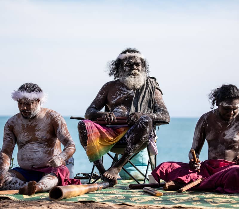 Three Aboriginal men with white body paint sitting on a mat by the ocean; the central man has a long white beard and a white feather headband.