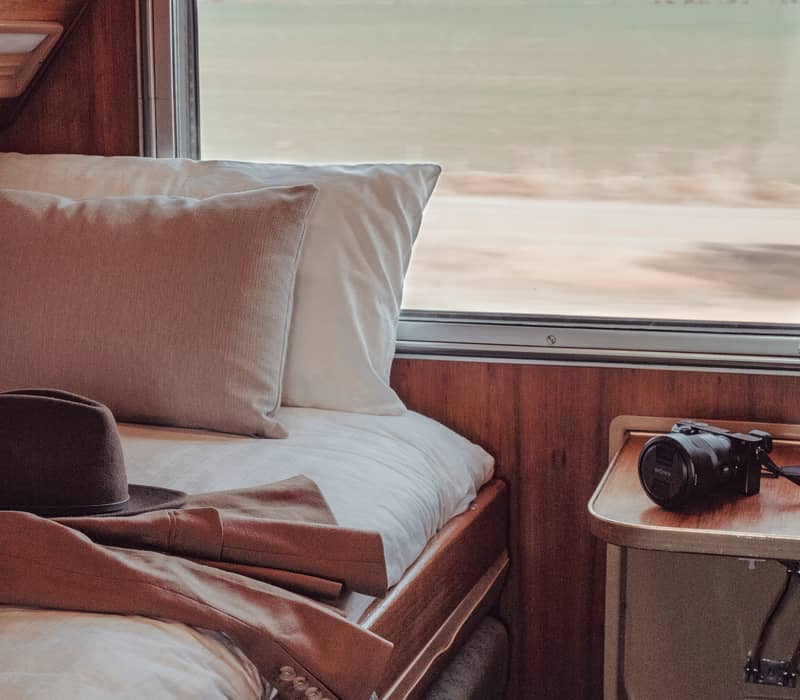 Interior of a cozy Gold Service train cabin with a bed, a camera on a bedside shelf, and a view of green rolling hills through the window.