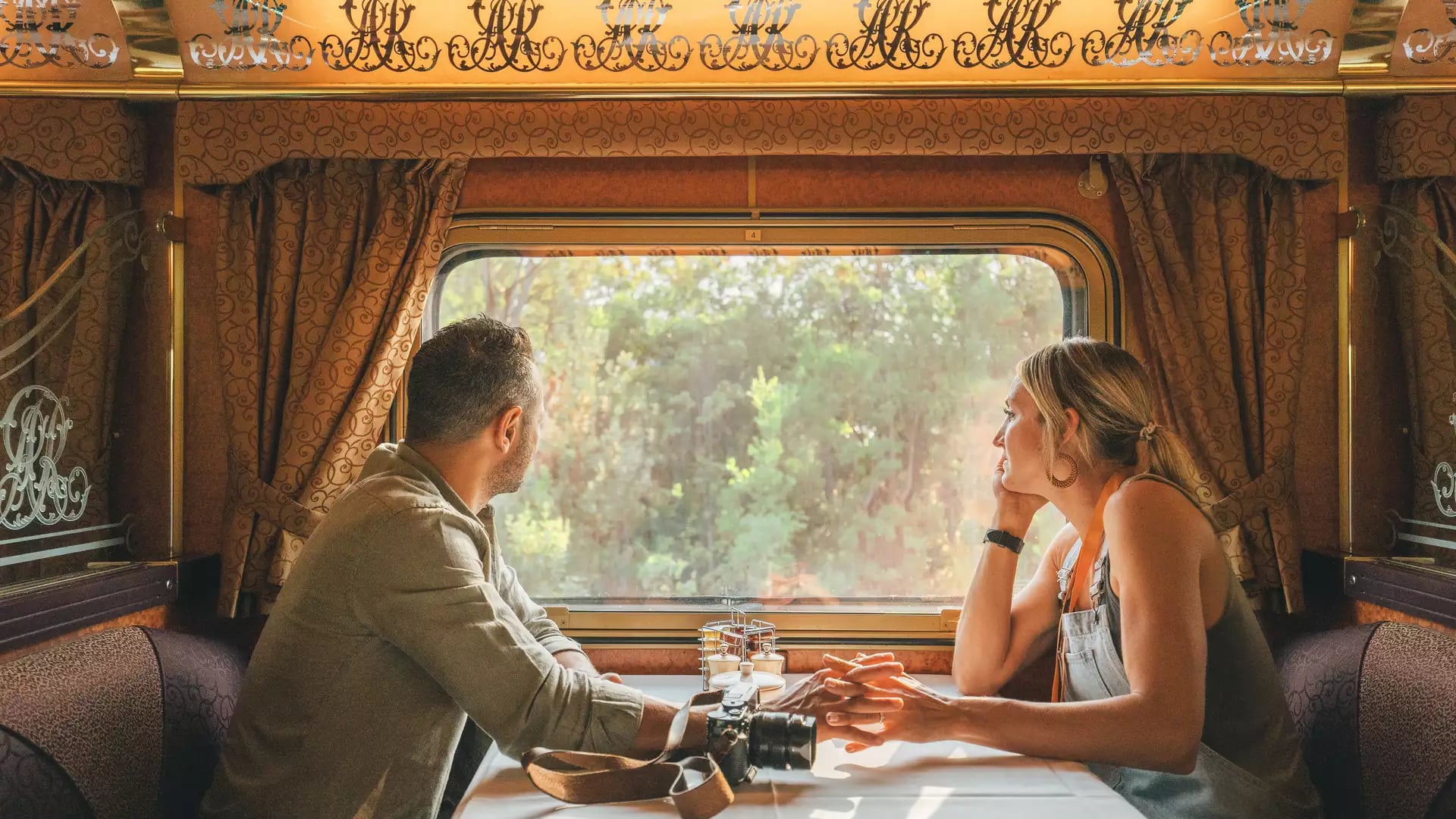 Interior view of a Gold Service Lounge on a Journey Beyond rail journey. A man and woman sitting at a white-clothed dining table inside a luxury Journey Beyond train carriage, looking out the window at green bushland.