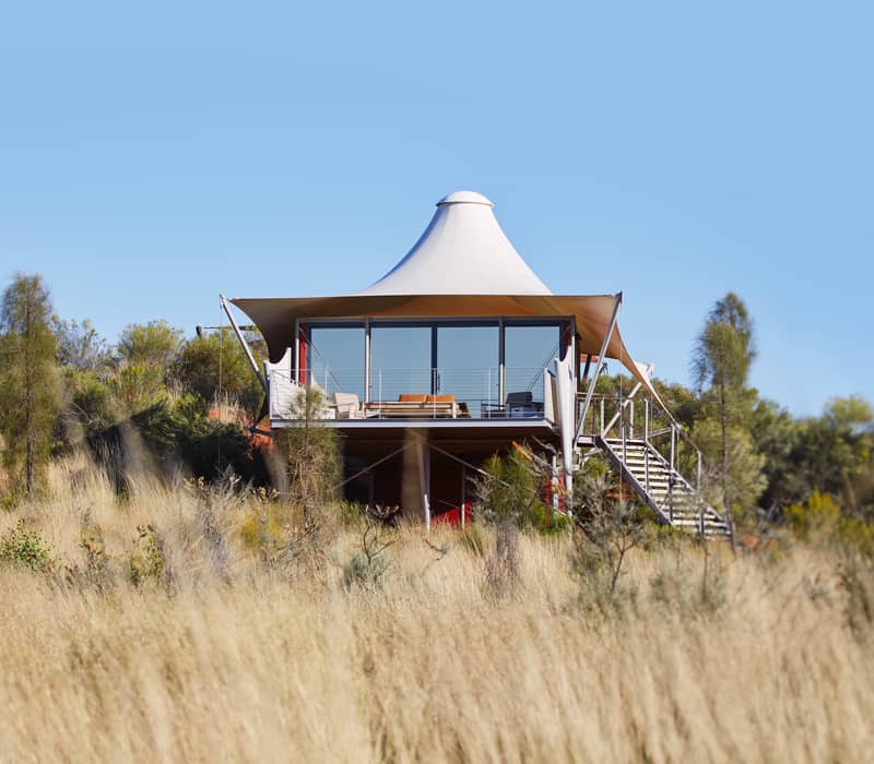A low-angle exterior view of a luxury elevated tent at Longitude 131°, featuring a distinctive white peaked canopy and floor-to-ceiling glass walls that provide panoramic views of the desert.