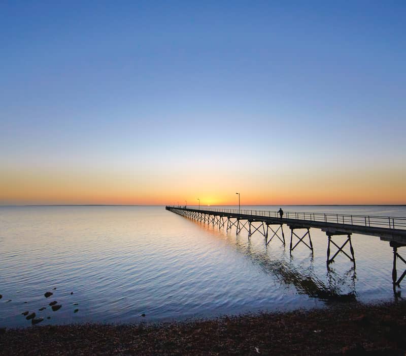 A long, silhouetted wooden Ceduna Jetty stretching out into a calm ocean toward a colorful orange and blue sunset horizon.