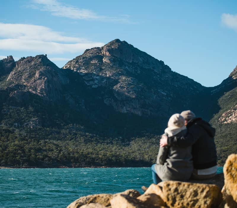 A man and woman sitting on a rock by the ocean, seen from behind, looking out at a large rocky mountain across blue water.