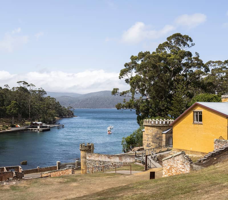 Stone ruins and a bright yellow house on a grassy hill overlooking a blue bay with small boats.