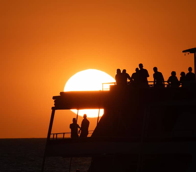 Silhouetted passengers on the deck of a cruise boat watch a massive golden sun dip below the horizon of the Arafura Sea.