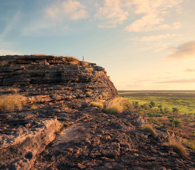 A wide-angle landscape shot from the rocky summit of Ubirr, showing visitors silhouetted against a golden sky overlooking the vast Nadab Floodplain in Kakadu National Park.