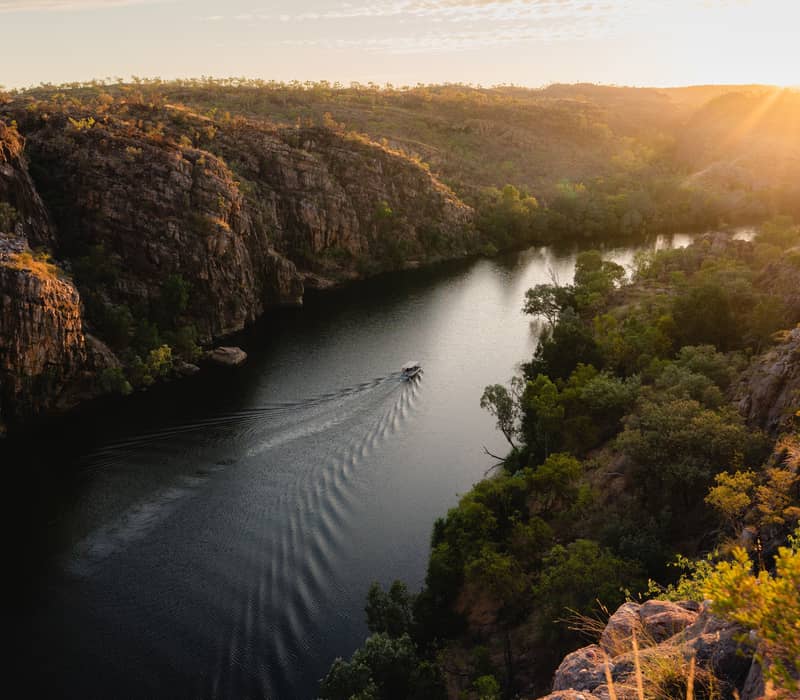 A high-angle view of a tour boat navigating the Katherine River as the sun sets over the Nitmiluk National Park escarpment, casting a brilliant golden light across the ancient landscape.