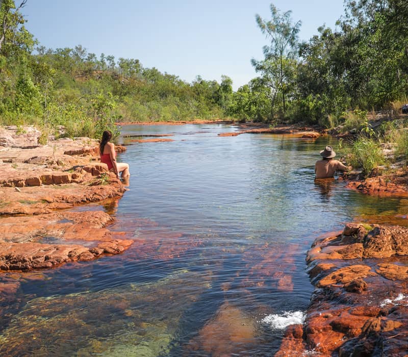 Two swimmers relax in a natural, crystal-clear infinity pool at the Upper Cascades, overlooking the vast tropical savanna of Litchfield National Park.