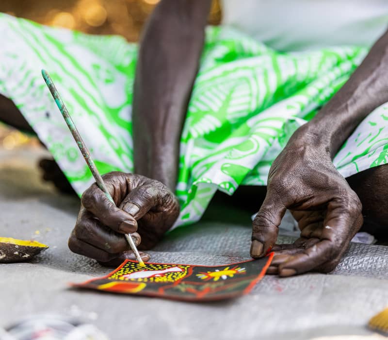 A close-up of a Tiwi artist's hands meticulously applying yellow ochre dots to a traditional bark-style painting.