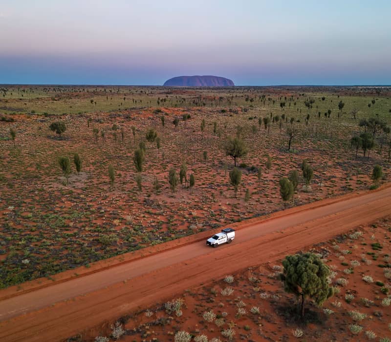 A white 4WD vehicle travels along a remote red dirt track through the vast desert landscape of Uluru-Kata Tjuta National Park, with the iconic silhouette of Uluru visible on the horizon.