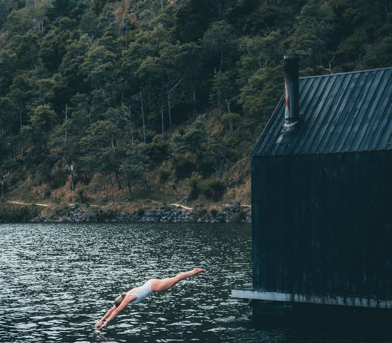 A person captured mid-dive into a deep, still lake next to a floating black timber cabin. A thin trail of white smoke rises from the cabin's chimney into the air, and a lush green forest hill rises steeply from the water's edge in the background.