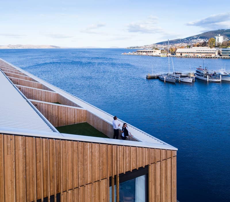 A luxurious Superior Waterfront Room at the MACq 01 Hotel on Hobart's waterfront, featuring a private balcony with views of the Derwent River and a large-scale mural headboard illustrating a story from Tasmania's history.