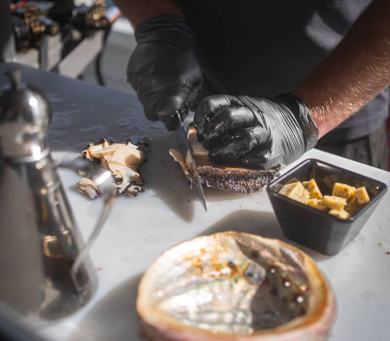 A chef expertly slices fresh Tasmanian abalone aboard a Tasmanian Wild Seafood Adventures vessel during a Sea to Sky Night Cruise, showcasing the region's premium local produce.