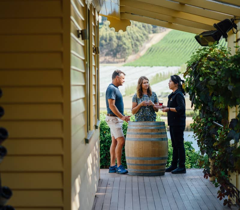 Under the shade of a yellow heritage building's eaves, three people stand on a wooden deck around a large oak wine barrel used as a table. A staff member is talking to a man and woman holding glasses of red wine, with a vast, sloping green vineyard visibl