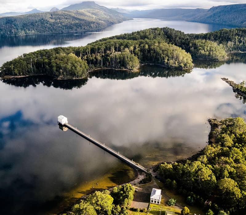 An expansive aerial view of a deep blue lake reflecting a cloudy sky, with a very long, straight pier leading to a small white building in the middle of the water. A lush green forested peninsula extends into the lake, and distant blue mountains line the horizon.