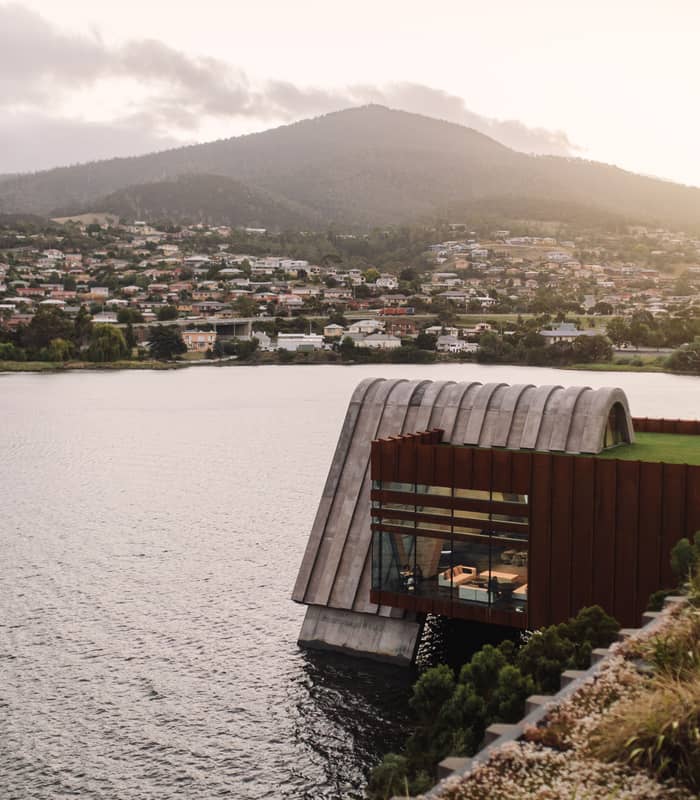 An eye-level shot across a body of water toward a modern building with a rounded concrete roof and a glass-walled lower section overlooking the river. In the background, rolling green hills are dotted with suburban houses under an overcast sky.