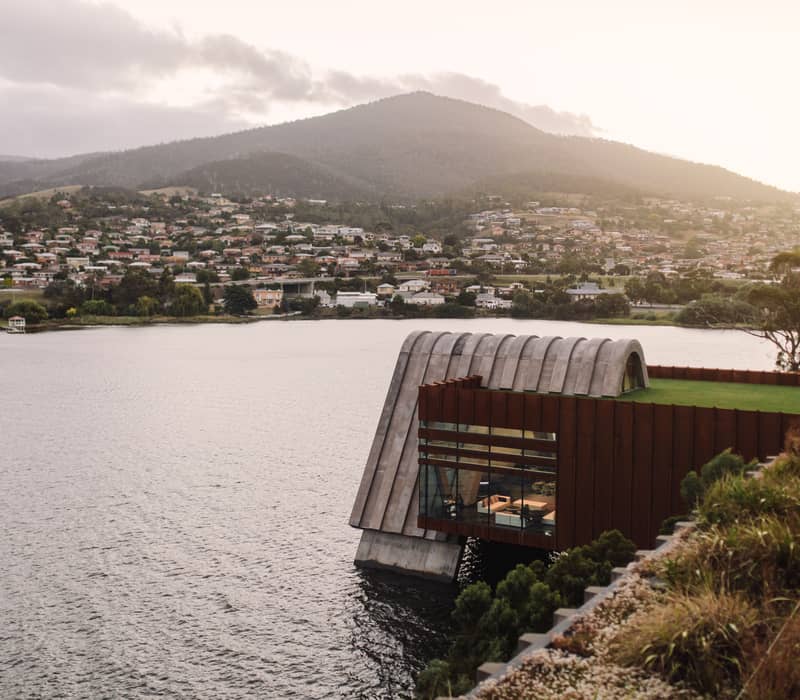 An eye-level shot across a body of water toward a modern building with a rounded concrete roof and a glass-walled lower section overlooking the river. In the background, rolling green hills are dotted with suburban houses under an overcast sky.