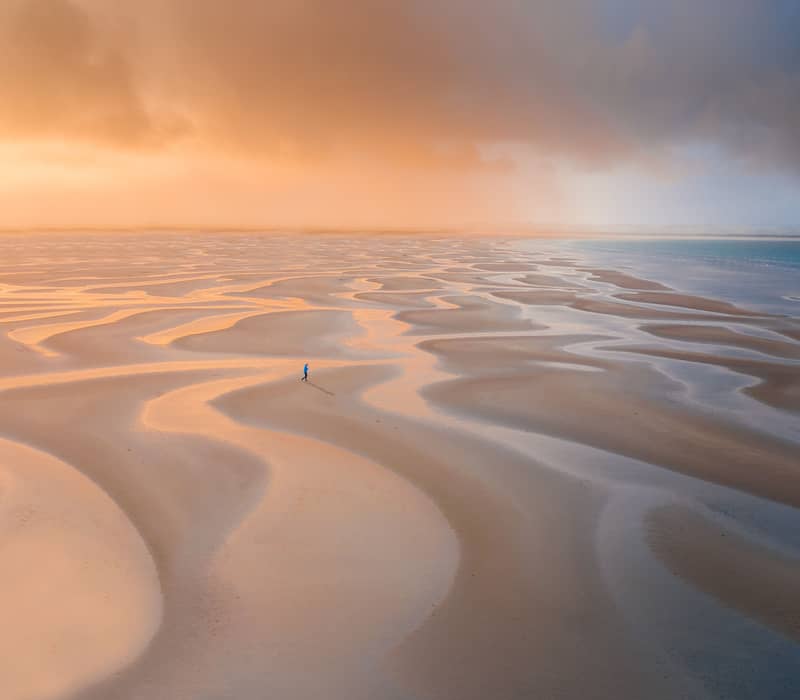 Aerial view of a lone person walking on vast, winding sandbanks illuminated by golden sunset light.