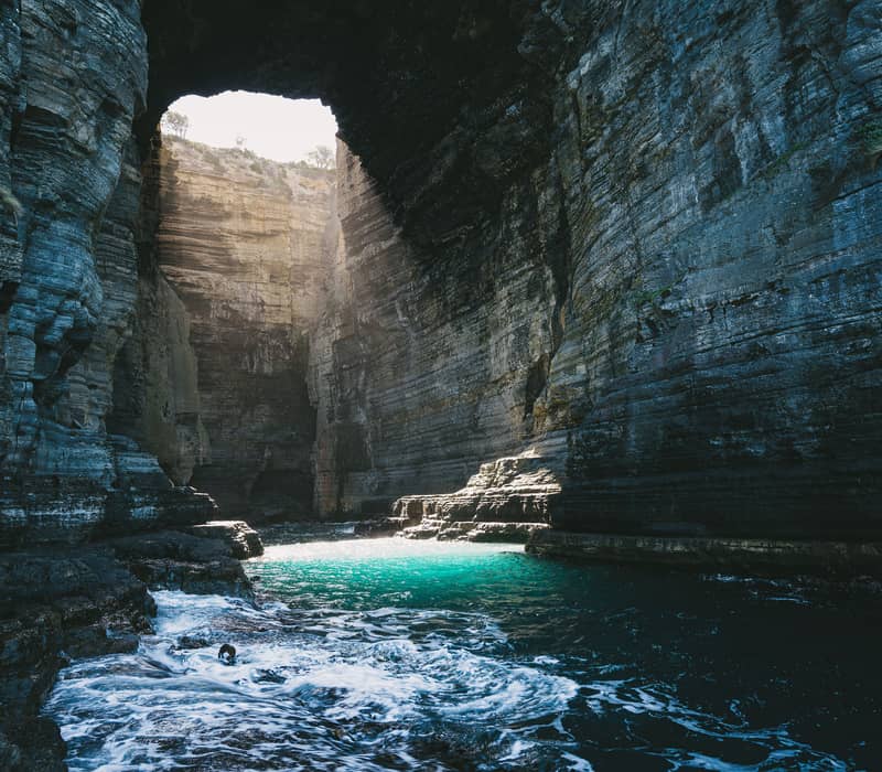 Sunlight streams through the opening of Tasman Arch, a massive natural bridge carved by the Southern Ocean into the high dolerite cliffs of the Tasman National Park. Located near Eaglehawk Neck, this geological marvel is part of a series of dramatic coastal formations on the Tasman Peninsula.