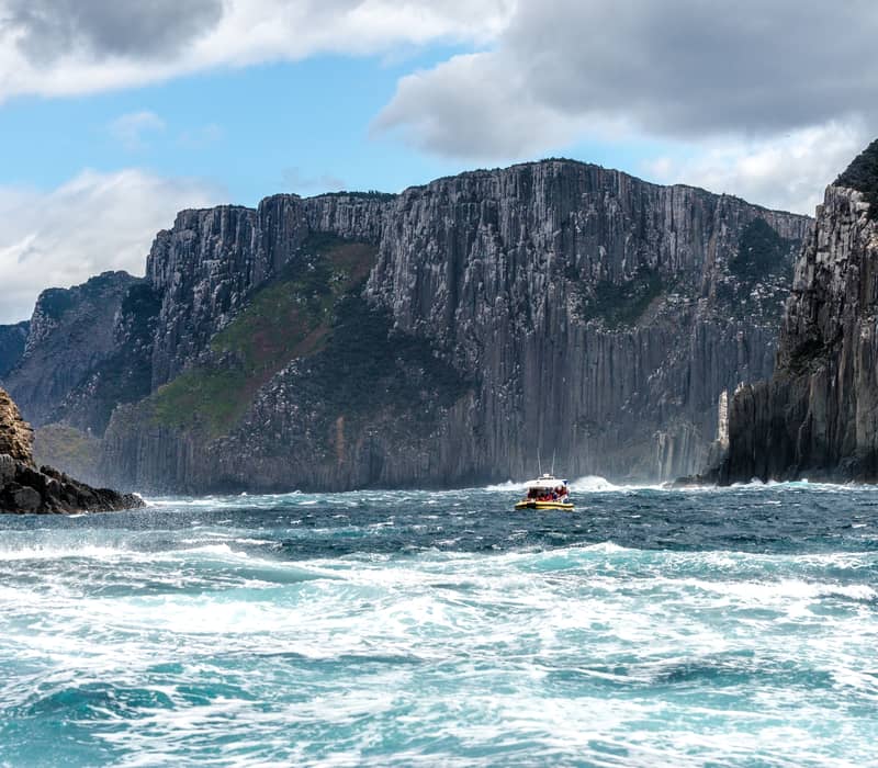 A yellow Pennicott Wilderness Journeys eco-cruise vessel navigates the surging swell beneath the world's highest vertical sea cliffs at Tasman Island on the Turrakana / Tasman Peninsula.
