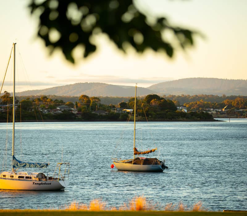 Two sailboats, one named "Tanglefoot," anchored on the calm blue waters of the Tamar River during a golden sunset, with the rolling hills and houses of the Tamar Valley in the background.