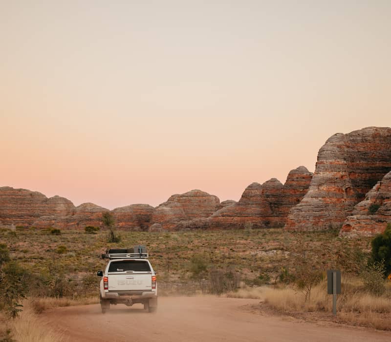 A rear view of a white 4WD kicking up dust on a dirt track. In the background, large striped sandstone domes rise under a dramatic red and orange sunset sky.