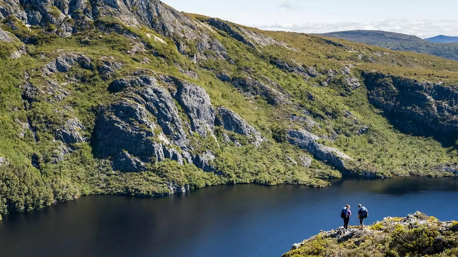 Two hikers standing on a rocky outcrop overlooking a lake and mountainous landscape.