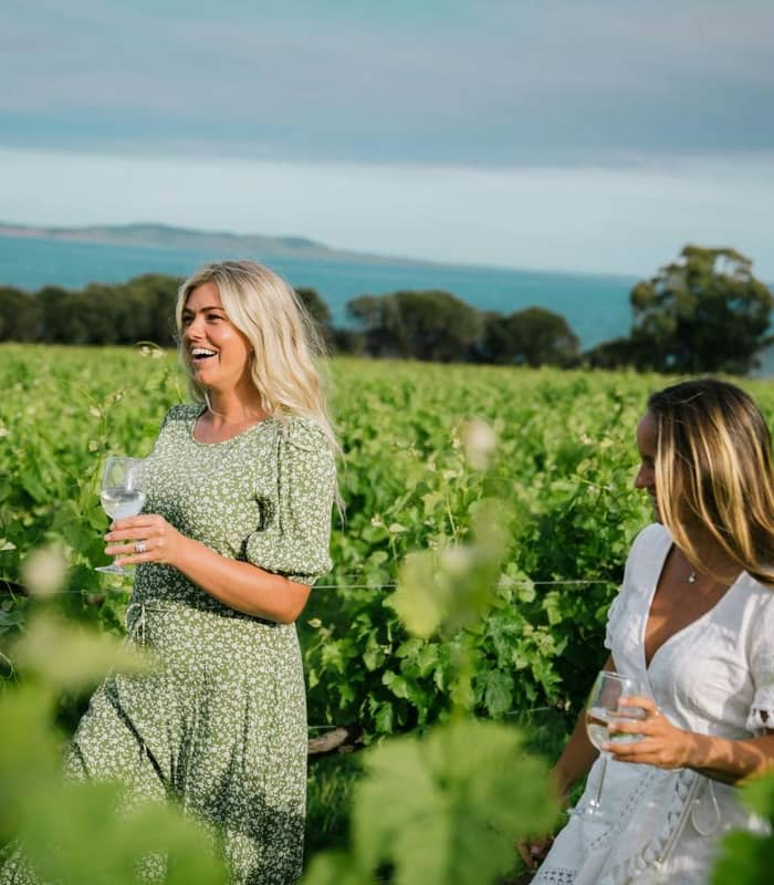 Two women holding wine glasses walk through a green vineyard with water and hills in the background.